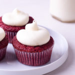 Straight on view of a plate of red velvet cupcakes with cream cheese frosting with a jug of milk in the background.