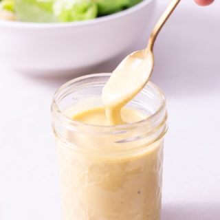 Straight on shot of a jar of Caesar Dressing with a spoon in it with Caesar Salad in the background.