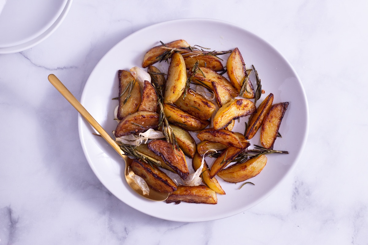 Overhead view of potato wedges with rosemary on a white platter with a gold spoon on a marble surface next to white serving plates.