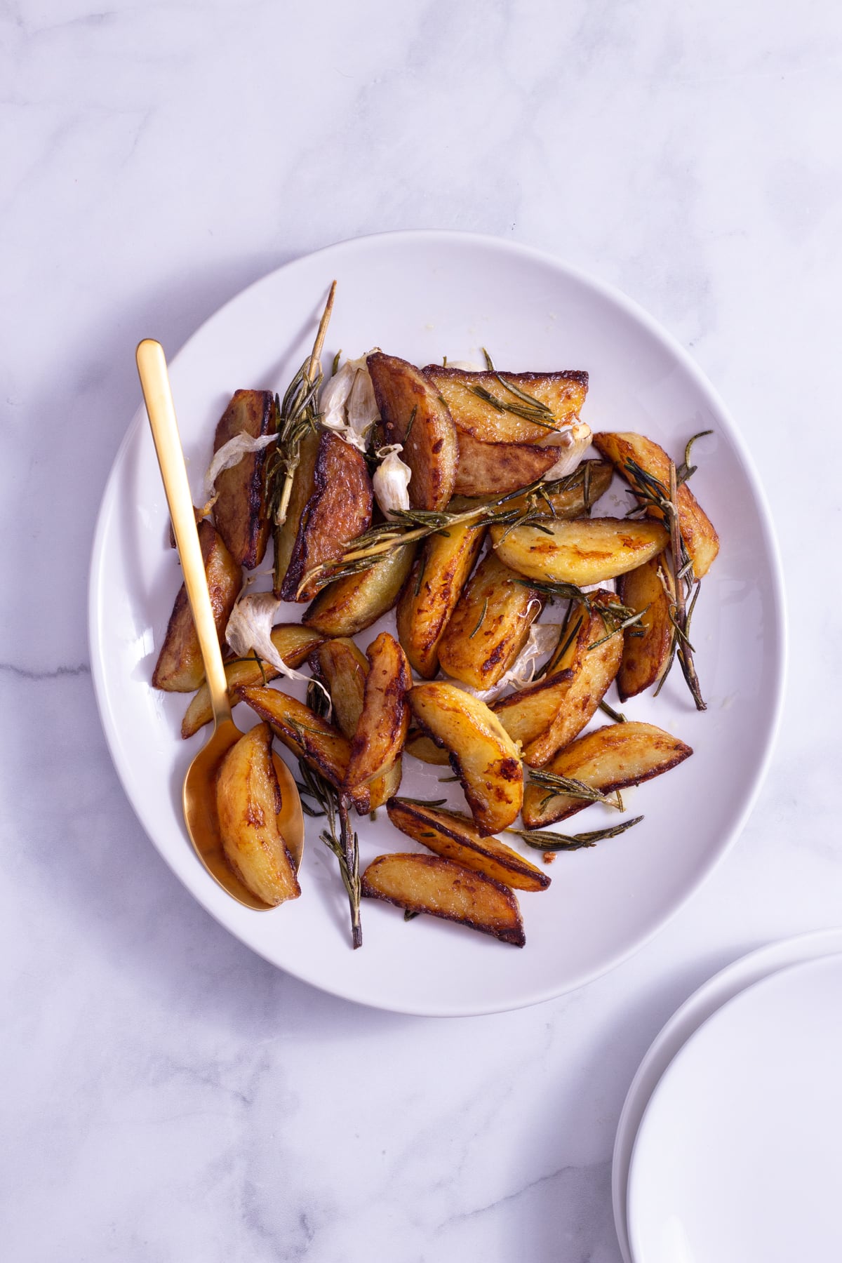 Overhead view of potato wedges with fresh rosemary and garlic cloves on a white platter with a gold spoon on a marble surface.