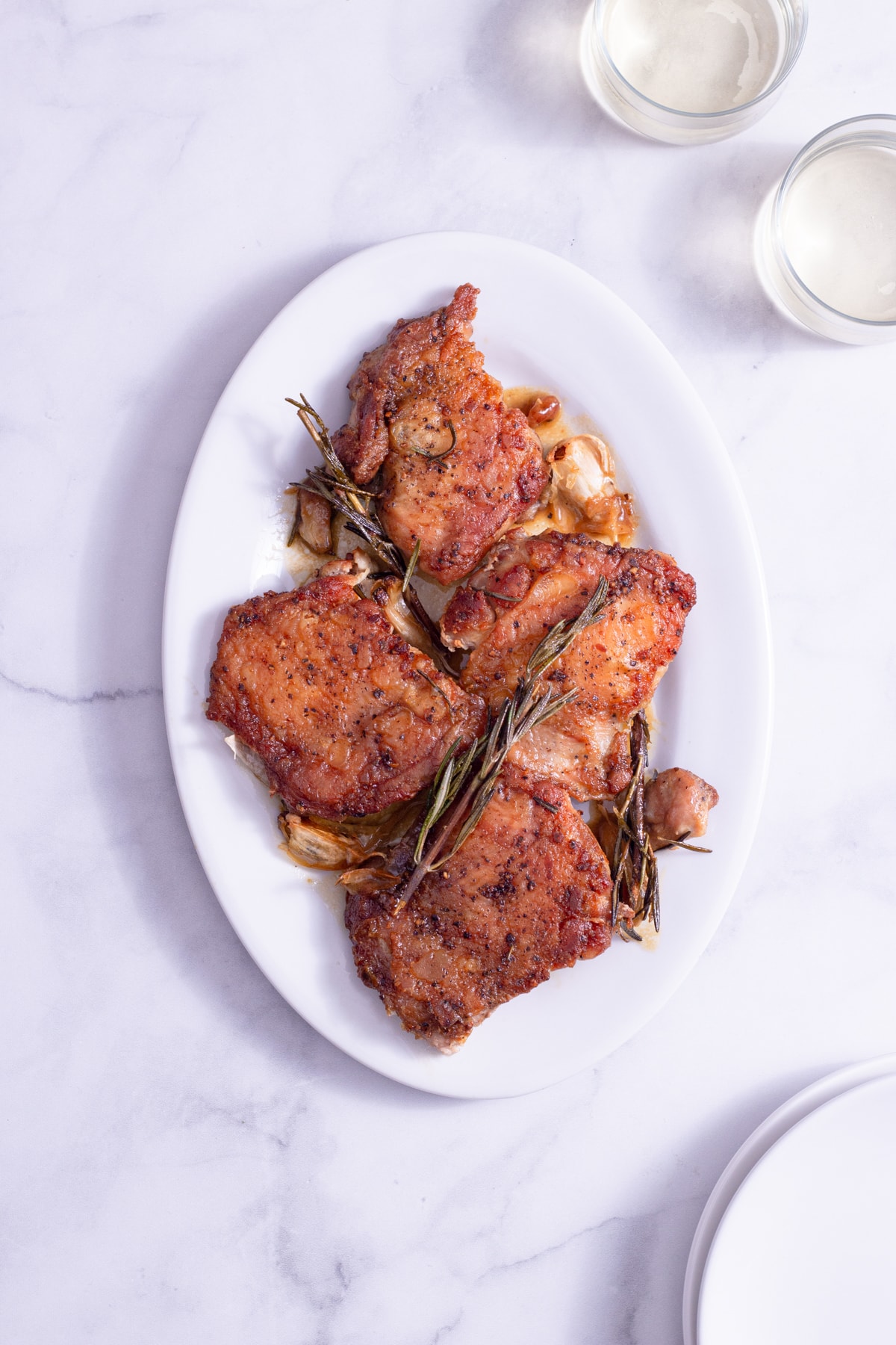 Overhead shot of a platter of Crispy Chicken Thighs with Rosemary and Garlic surrounded by glasses of wine and white plates on a marble surface.