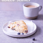 Angled view of a lemon blueberry scone on a plate surrounded by scattered fresh blueberries with a mug of coffee in the background on a light grey surface.