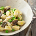 Angled view of a bowl of Homemade Gnocchi with Morel Mushrooms, Peas and Fava Beans on a light wood surface.