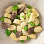 Overhead, close up view of a bowl of Homemade Gnocchi with Morel Mushrooms, Peas and Fava Beans on a light wood surface.