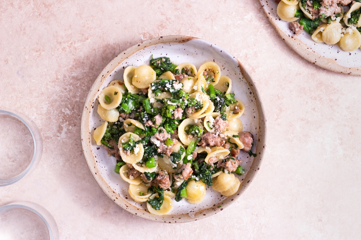 Overhead view of a bowl of orecchiette with Sausage and Broccoli Rabe surrounded by a plate of pasta and a glass on a light brown, textured surface.