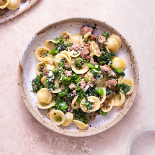 Overhead view of a bowl of Pasta with Sausage and Broccoli Rabe surrounded by a plate of pasta and two glasses on a light brown, textured surface.