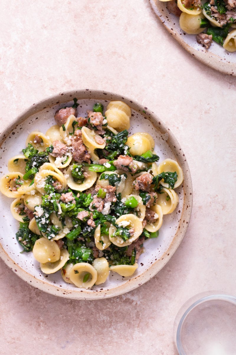 Overhead view of a bowl of Pasta with Sausage and Broccoli Rabe surrounded by a plate of pasta and a glass on a light brown, textured surface.