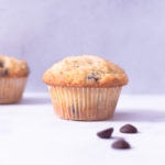 Close up, straight on view of a muffin with chocolate chips on a white surface with a light blue background.