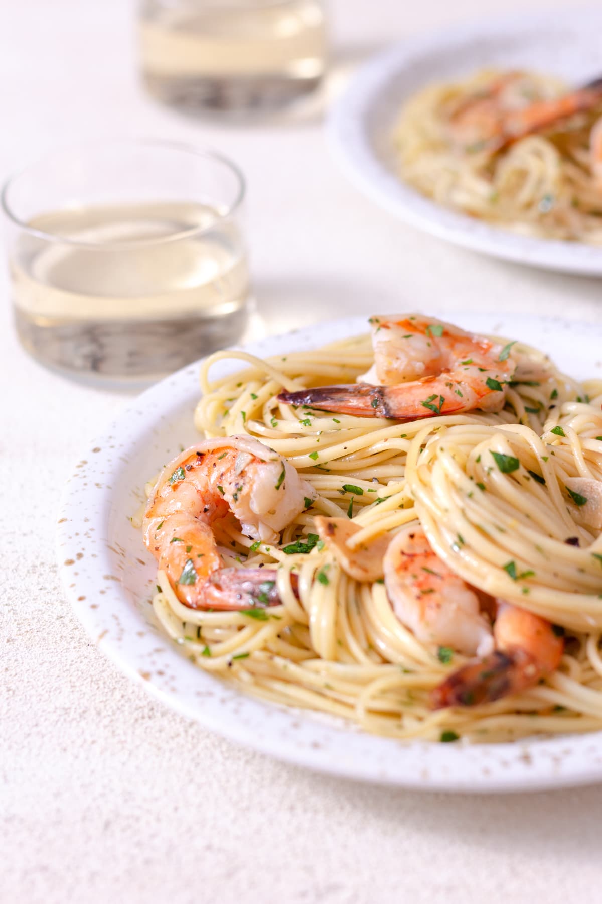 Angled view of a plate of shrimp pasta with wine glasses and another plate in the background on a white plaster surface.