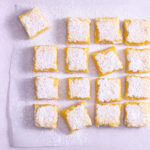 Overhead view of lemon bars on parchment on a light surface.
