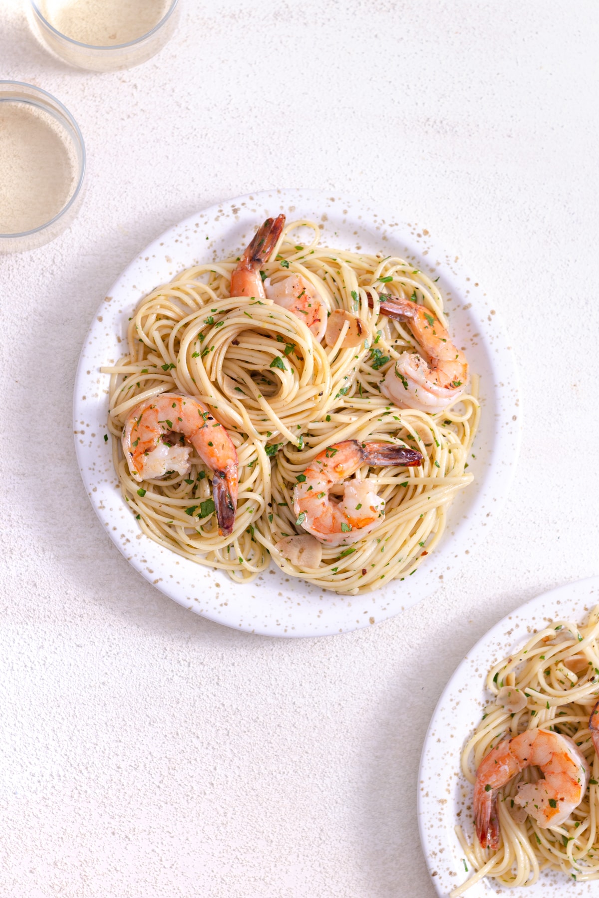 Overhead view of a plate of lemon garlic shrimp pasta next to glasses of wine on a white plaster surface.