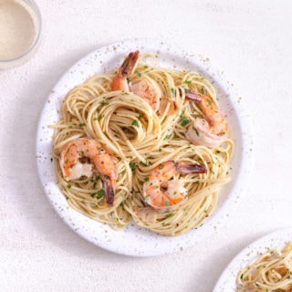 Overhead view of a plate of shrimp pasta surrounded by a wine glass on a white plaster surface.