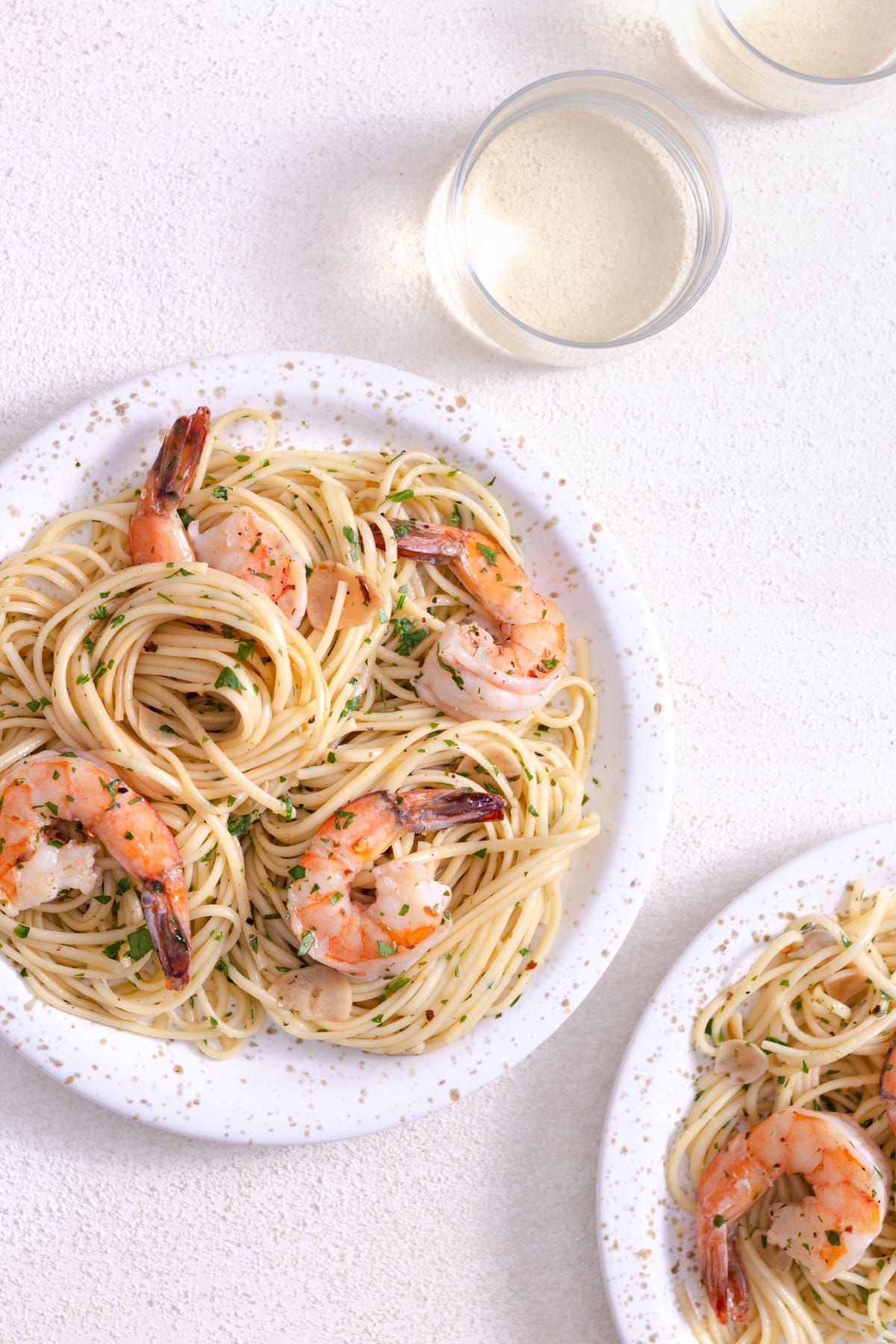 Overhead view of two plates of spaghetti with shrimp next to glasses of wine on a white plaster surface.