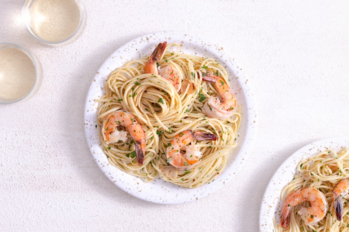 Overhead view of two plates of shrimp spaghetti next to glasses of wine on a white plaster surface.