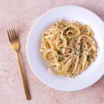 Overhead, close up view of a plate of Fettuccine Alfredo garnished with parsley surrounded by a copper fork and a bowl of grated parmesan cheese on a light brown, textured surface.