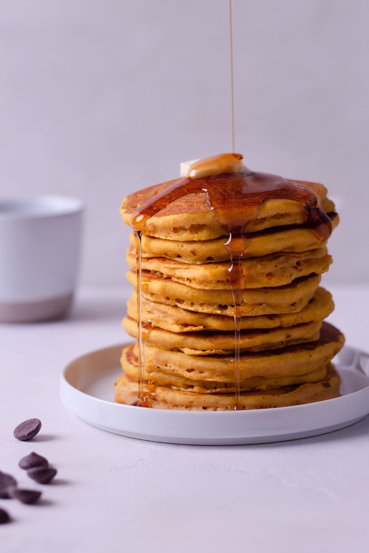 Straight on shot of a high stack of pumpkin chocolate chip pancakes with butter on top and maple syrup dripping down, with a coffee mug in the background.