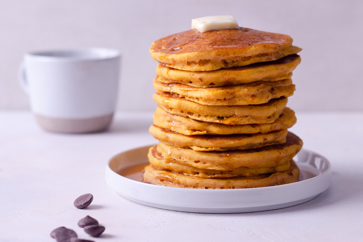 Straight on view of a stack of pumpkin pancakes on a plate, surrounded by scattered chocolate chips with a coffee mug in the background.
