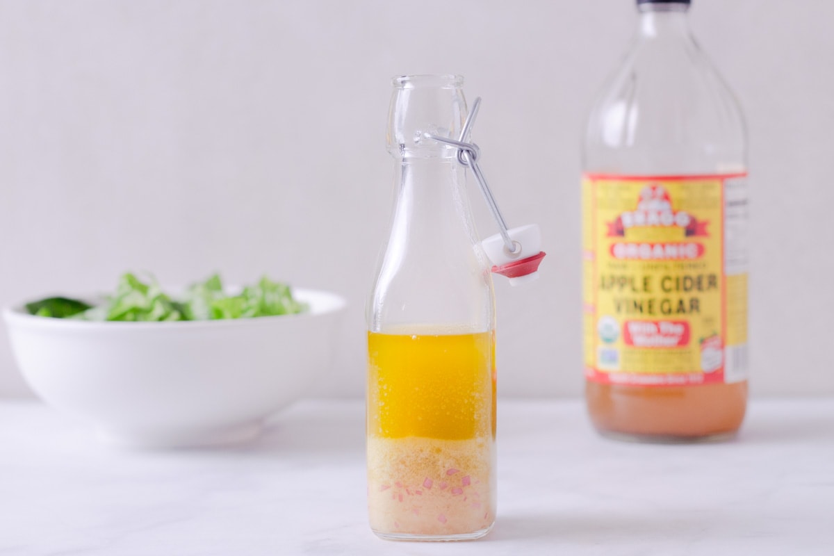 Straight on view of salad dressing in a glass container with a salad and bottle of vinegar in the background.