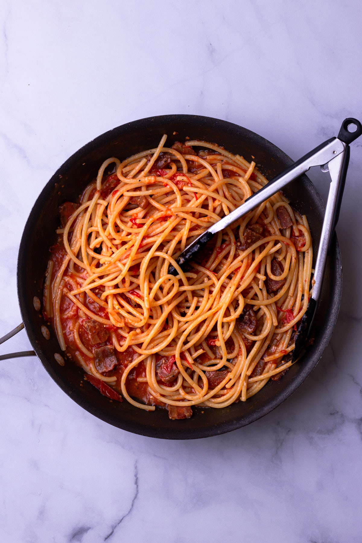 Overhead view of a pan with pasta and tomato-guanciale sauce with tongs on a marble surface.