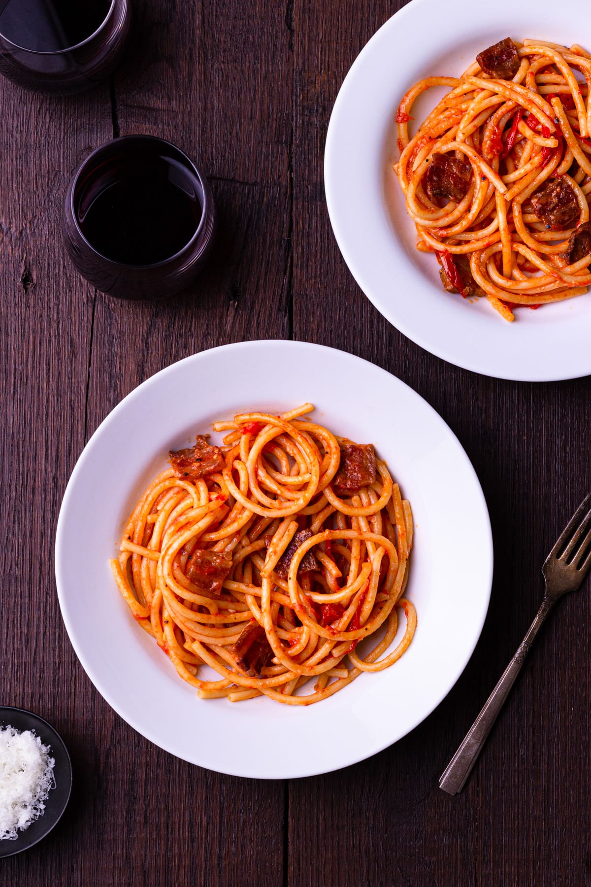 Overhead view of two bowls of Bucatini All'Amatriciana on a dark wood surface surrounded by glasses of red wine a fork.