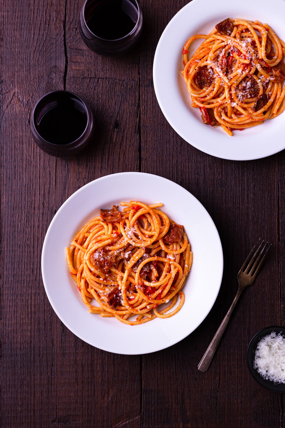 Two bowls of pasta with Amatriciana sauce topped with grated pecorino cheese, surrounded by glasses of red wine and a fork.
