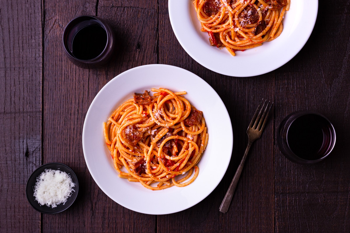 Overhead view of two bowls of pasta with fresh tomato sauce and guanciale surrounded by glasses of red wine and a bowl of pecorino cheese.