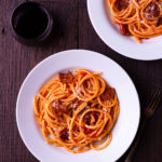 Overhead view of two bowls of pasta with tomato sauce and guanciale on a dark wood surface surrounded by wine glasses, grated cheese and a fork.