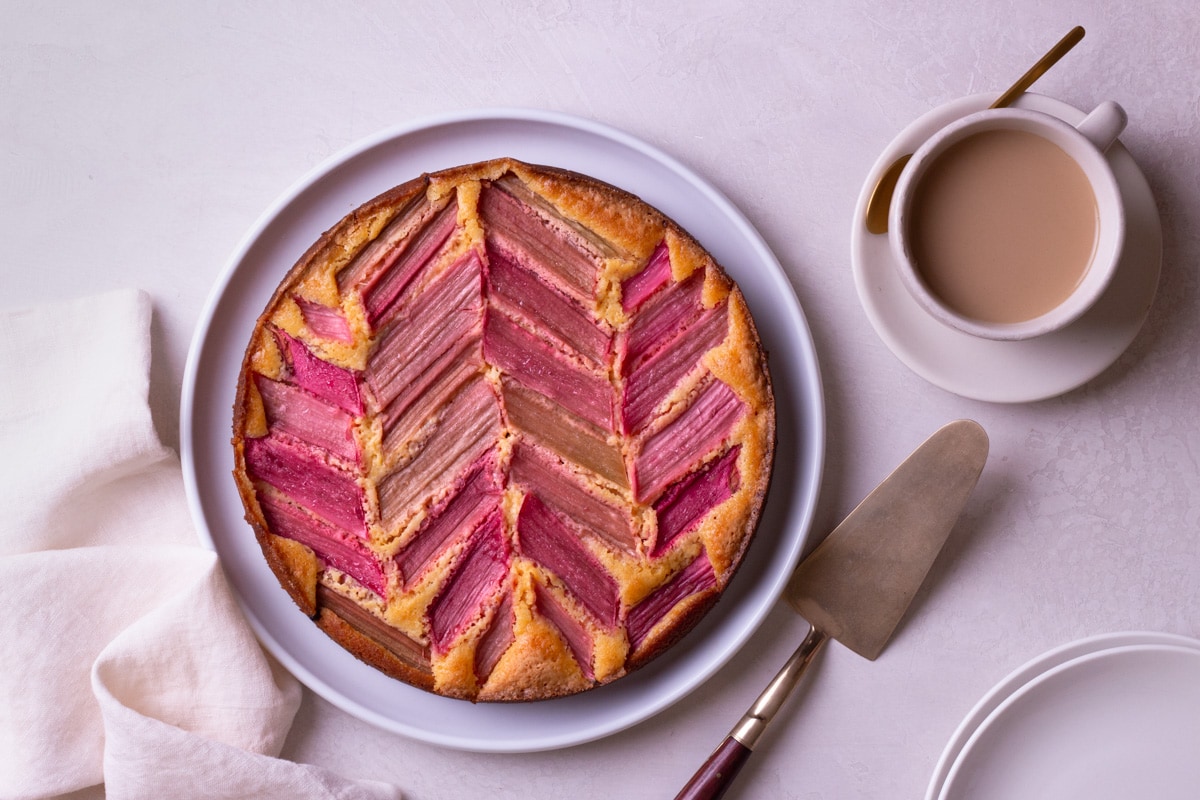 Overhead shot of a rhubarb cake on a cream surface surrounded by a cake knife, cup of coffee, plates and dish towel.