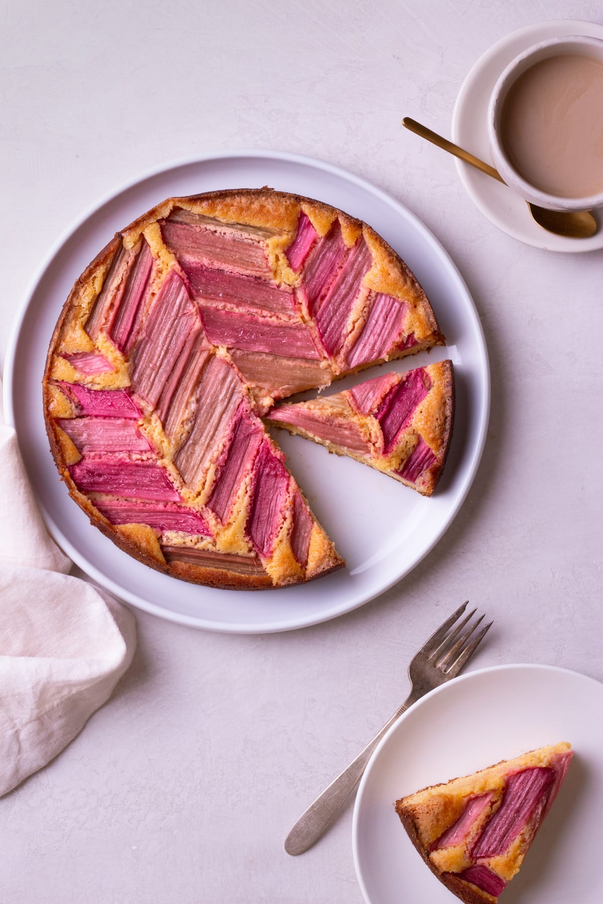 Overhead view of Rhubarb Custard Cake with slices cut out of it on a cream surface surrounded by a cup of coffee and a slice of cake.
