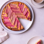 Overhead view of a rhubarb cake with slices cut out next to a plated slice and a cup of coffee.