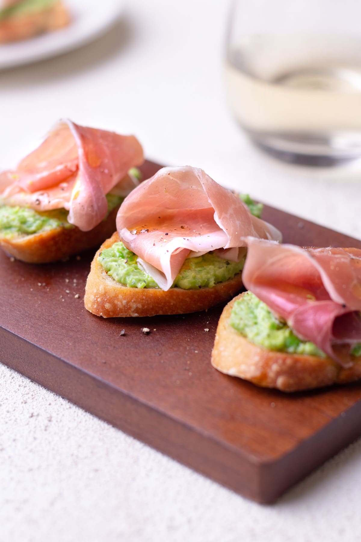Closeup straight on shoot of 3 crostini top with pea and ricotta puree and prosciutto on a cutting board.
