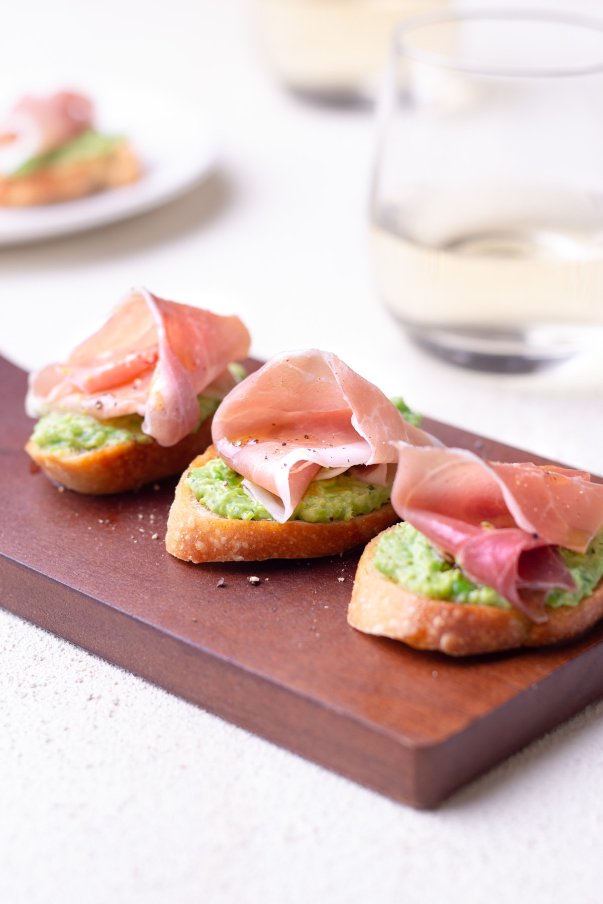Crostini topped with pea-ricotta spread and prosciutto on a cutting board with white wine and a plate in the background.