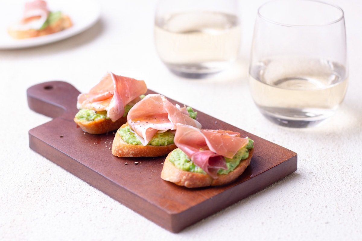 Pea and Ricotta Crostini topped with prosciutto on a small wood cutting board over a textured surface surrounded by glasses of white wine and a plate in the background.