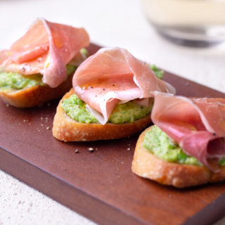 Closeup straight on shoot of 3 crostini top with pea and ricotta puree and prosciutto on a cutting board.