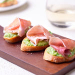 Crostini topped with pea-ricotta spread and prosciutto on a cutting board with white wine and a plate in the background.