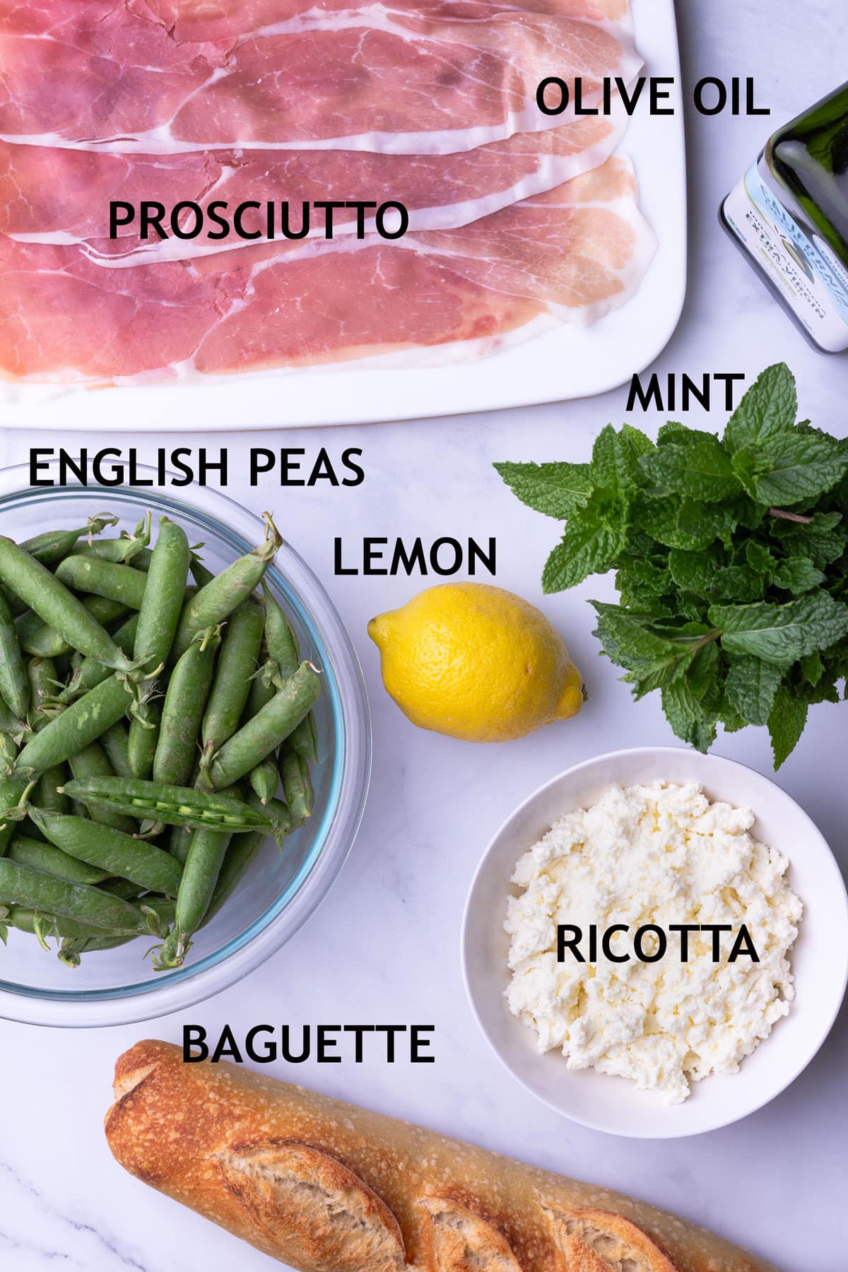 Overhead view of ingredients for crostini including prosciutto, olive oil, fresh English peas, lemon, mint, ricotta cheese and a baguette.