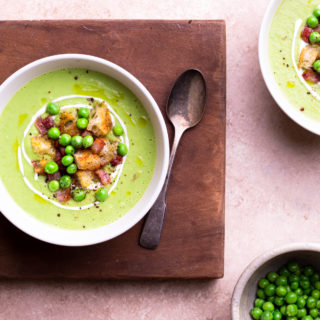 Overhead close up shot of a bowl of creamy green pea soup topped with pancetta and croutons on a wood cutting board over a light brown surface.