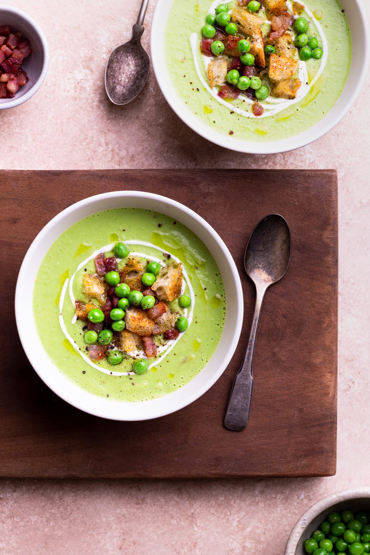 Two bowls of green pea soup on a wood cutting board over a light brown surface.