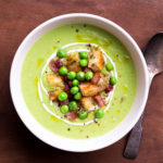 Closeup overhead view of a bowl of Green Pea Soup topped with yogurt, pancetta, peas and croutons.