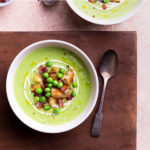 Overhead view of two bowls of creamy green pea soup topped with yogurt, peas, croutons and pancetta on a wood cutting board surrounded by spoons and bowls of toppings on a light brown surface.