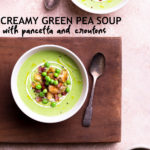 Overhead view of two bowls of creamy green pea soup topped with yogurt, peas, croutons and pancetta on a wood cutting board surrounded by spoons and bowls of toppings on a light brown surface.