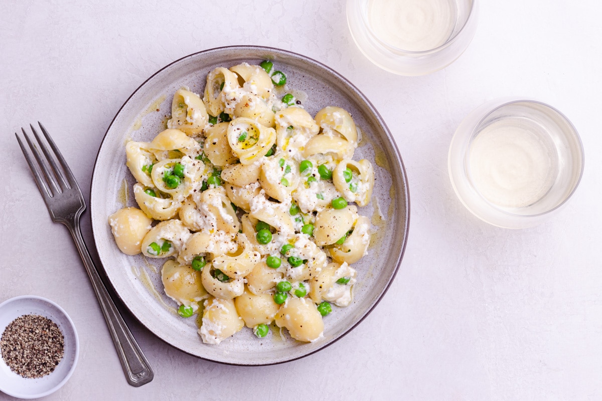 Overhead shot of a bowl of pasta and peas on a creamy surface with a fork and glasses of white wine.