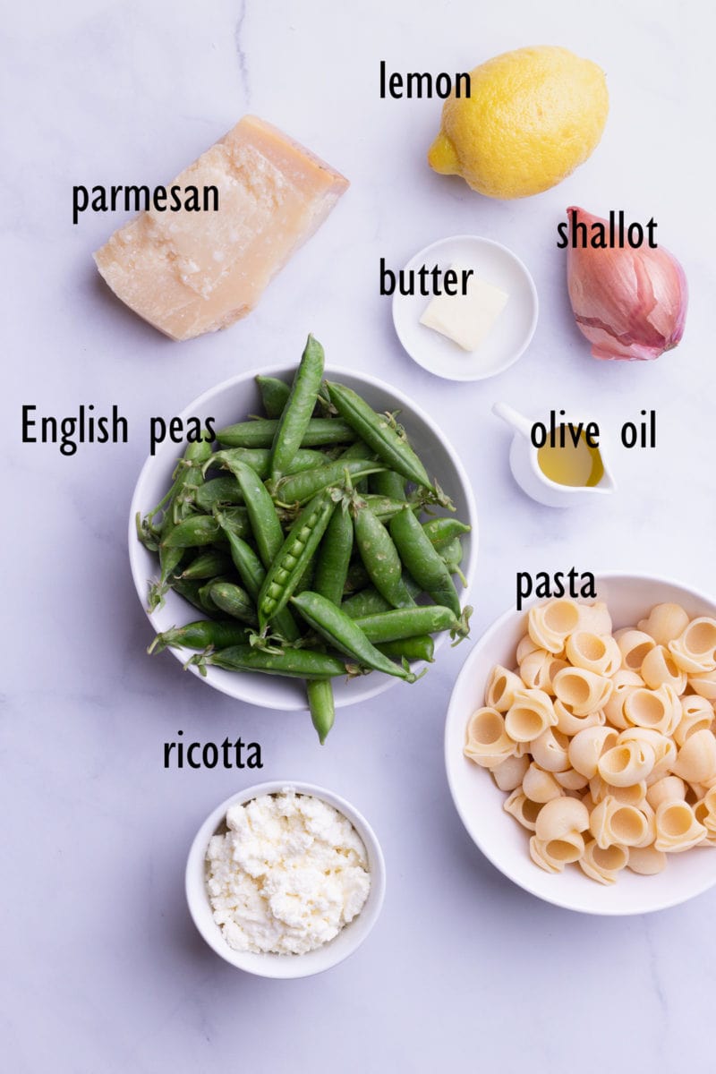 Overhead view of ingredients including English peas in the pod, butter, olive oil, a shallot, a lemon, ricotta and pasta.