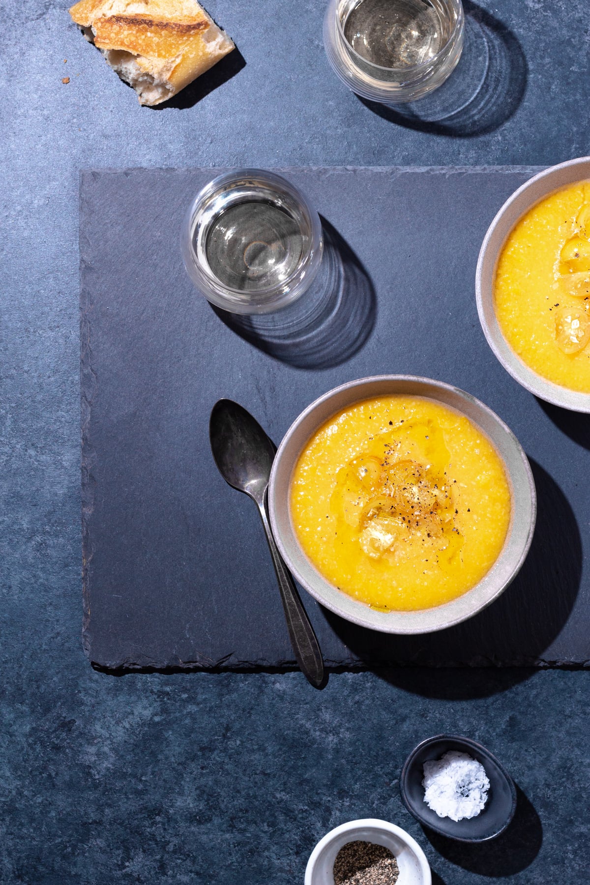 Overhead view of two bowls of yellow gazpacho and a grey-blue surface surrounded by glasses of white wine, crusty bread, a spoon and salt and pepper bowls.