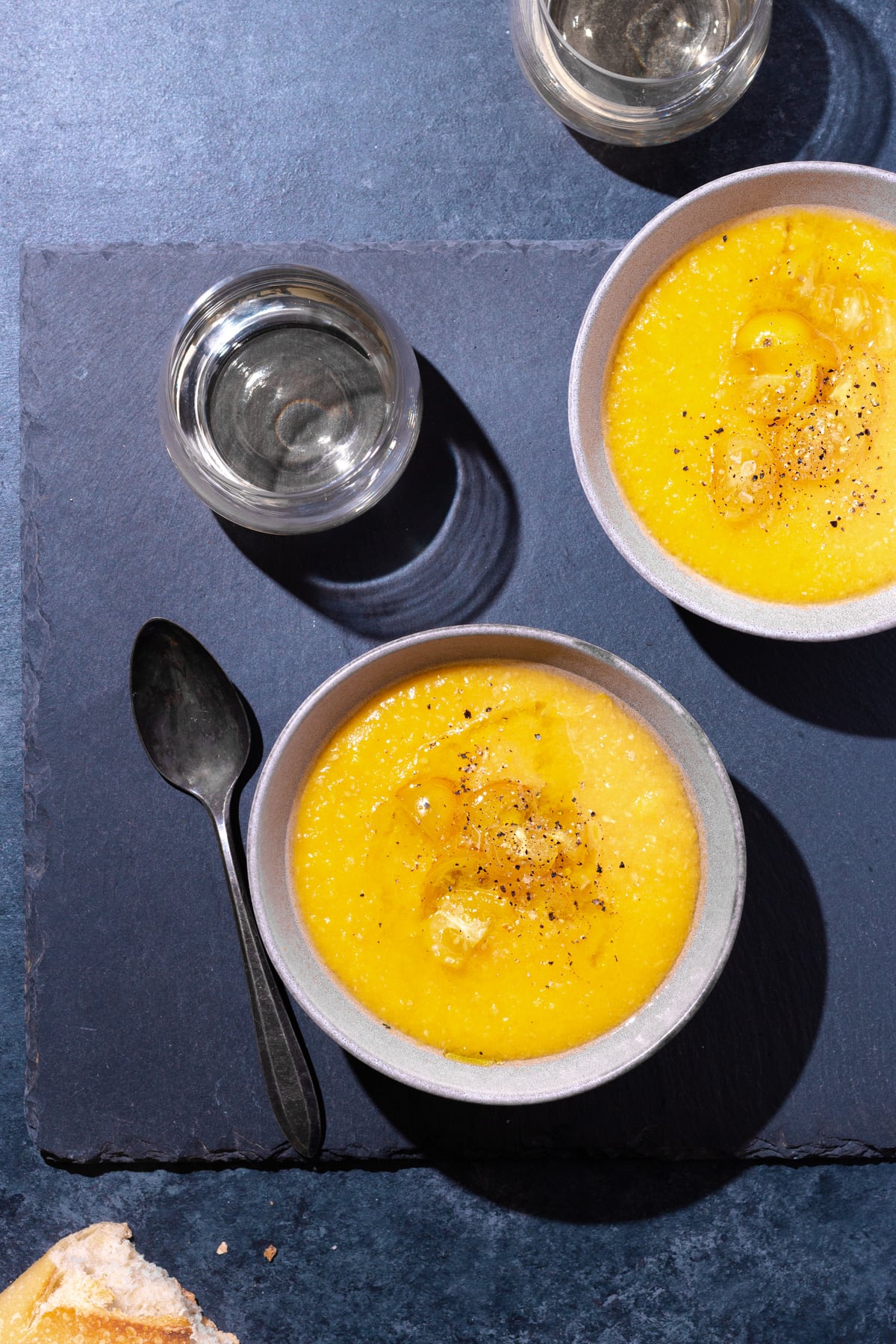 Overhead view of two bowls of yellow gazpacho on a slate surface next to wine glasses, a spoon and a piece of bread.