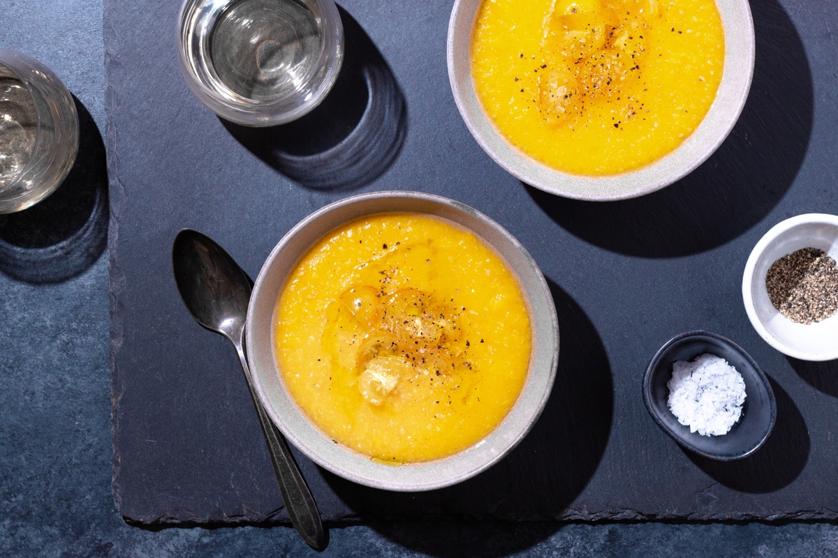 Overhead view of two bowls yellow tomato gazpacho surrounded by glasses of wine, salt and pepper pinch bowls, and a spoon on a slate surface.
