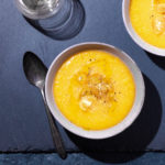 Overhead view of two bowls of yellow gazpacho on a slate surface next a wine glass and spoon.