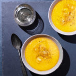Overhead view of two bowls of yellow gazpacho on a slate surface next to wine glasses, a spoon and a piece of bread.