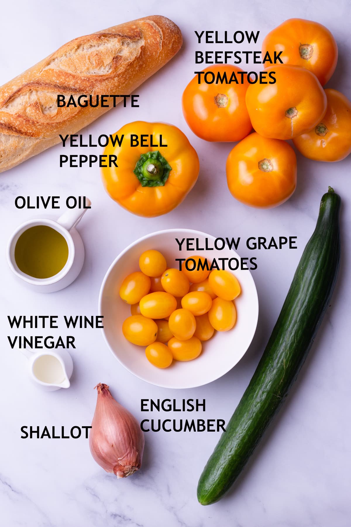 Overhead view of ingredients for gazpacho including bread, yellow tomatoes, a yellow bell pepper, an English cucumber, a shallot, olive oil, and white wine vinegar.