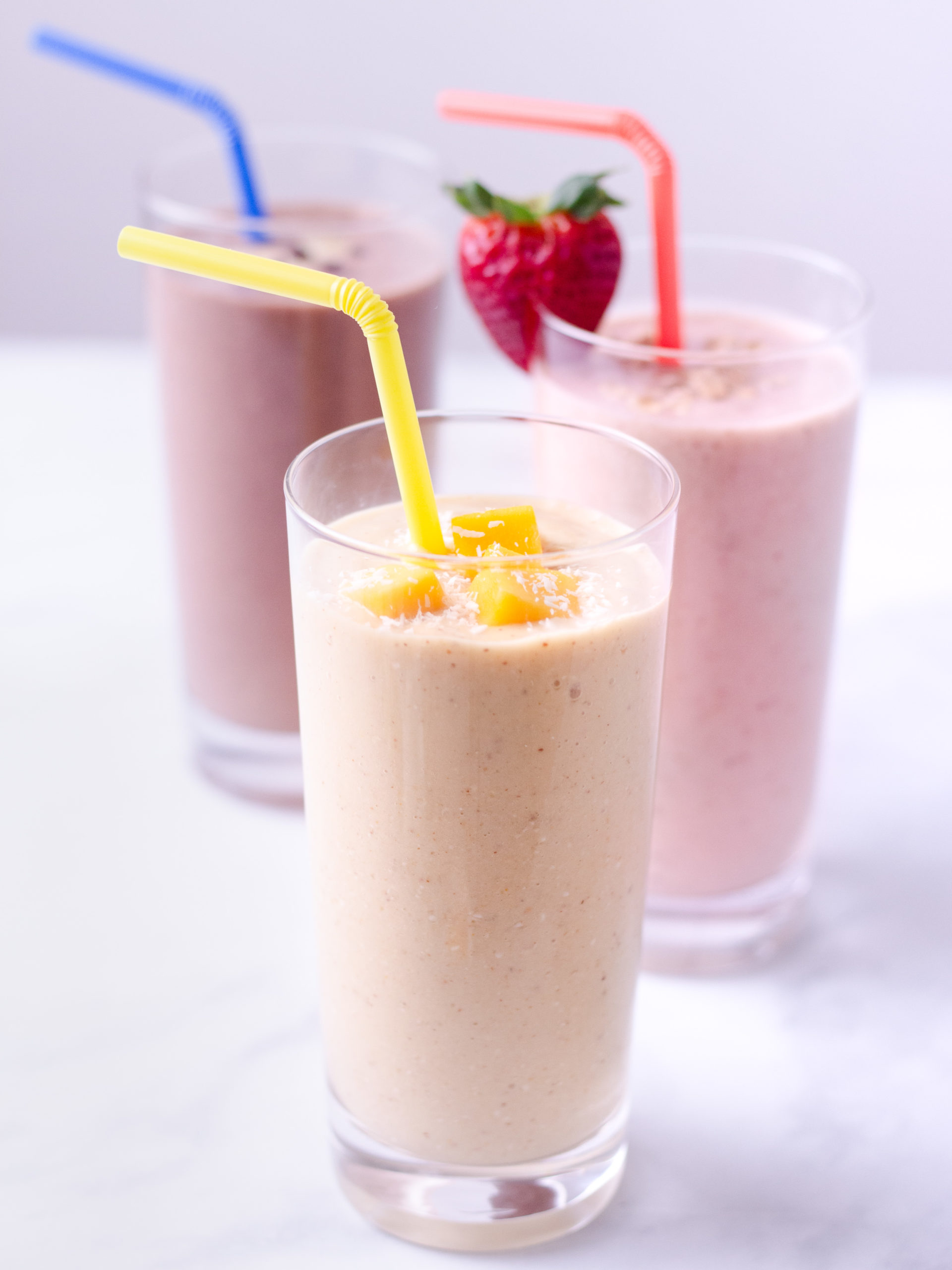 Three oat smoothies, including strawberry banana, turmeric mango and chocolate peanut butter straight on in glasses with straws on a marble surface.